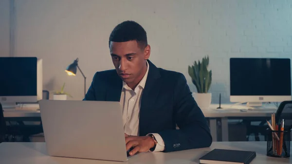 Hombre de negocios afroamericano usando portátil cerca de portátil en la oficina por la noche - foto de stock