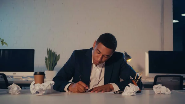 Empresario afroamericano escribiendo en cuaderno cerca de papel arrugado y café para llevar por la noche - foto de stock
