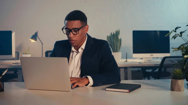 African american businessman in eyeglasses using laptop in office in evening — Stock Photo