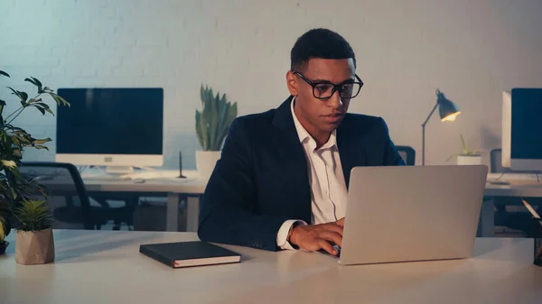 African american manager in eyeglasses using laptop near notebook in office at night — Stock Photo