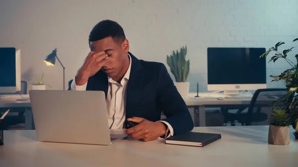 Uomo d'affari africano americano stanco seduto vicino a laptop e notebook in ufficio la sera — Foto stock
