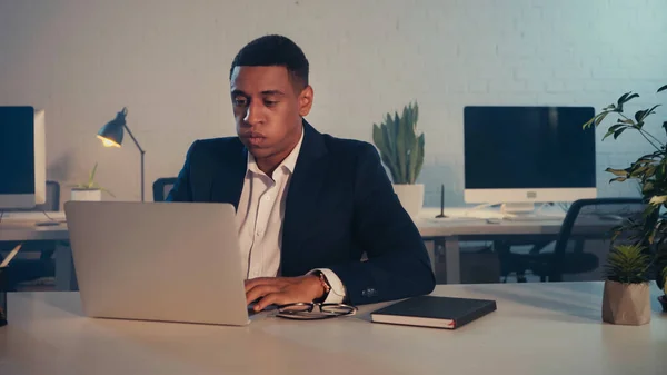 Виснажений афроамериканський менеджер використовує ноутбук біля окулярів і блокнот вночі — стокове фото