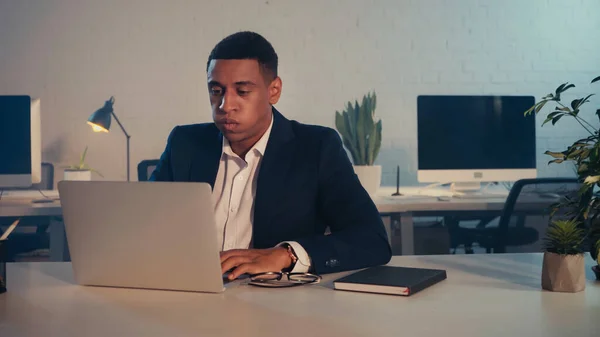 Exhausted african american manager using laptop near eyeglasses and notebook in office at night — Stock Photo