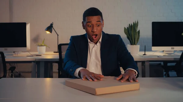 Excited african american businessman holding takeaway pizza in office at night — Stock Photo
