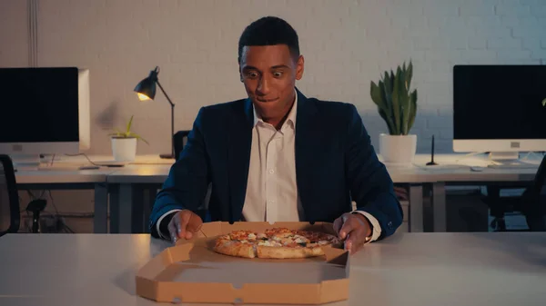Удовлетворенный африканский американский бизнесмен держит коробку с пиццей на вынос в офисе ночью — стоковое фото