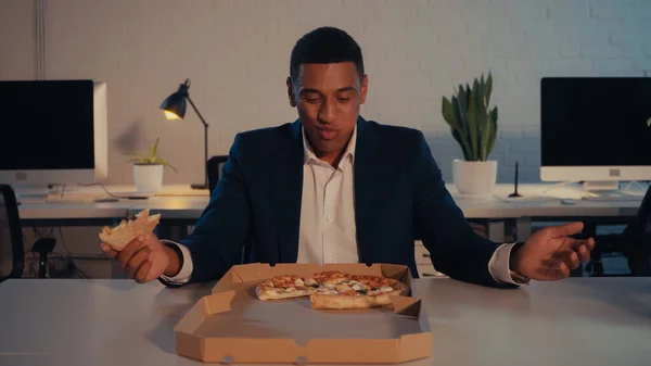 Joyeux homme d'affaires afro-américain tenant une pizza près de la boîte à emporter au bureau le soir — Photo de stock