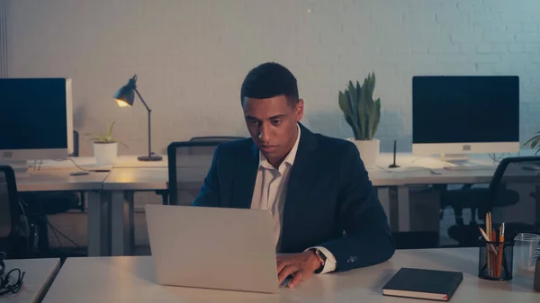 Gestionnaire afro-américain en costume à l'aide d'un ordinateur portable au bureau la nuit — Photo de stock