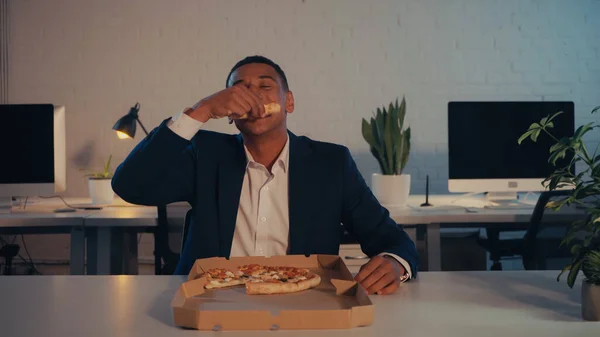 African american manager in suit eating takeaway pizza near box in office in evening — Stock Photo