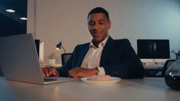 Посмішка афроамериканського бізнесмена, який дивиться на пончик біля кави і ноутбука в офісі. — стокове фото