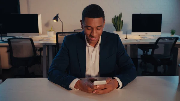 Negocio afroamericano positivo en traje usando smartphone en la oficina por la noche - foto de stock