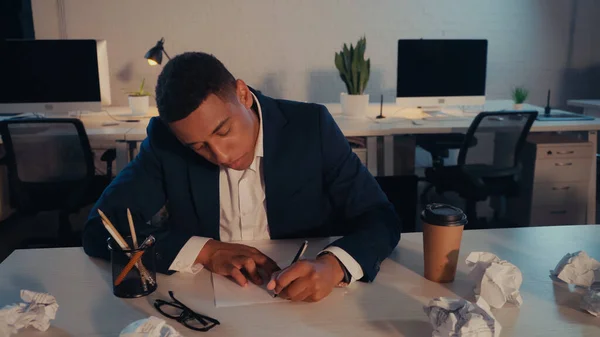 Afroamerikanischer Geschäftsmann schreibt an Dokument in der Nähe von zerknülltem Papier und Kaffee im Büro am Abend — Stockfoto