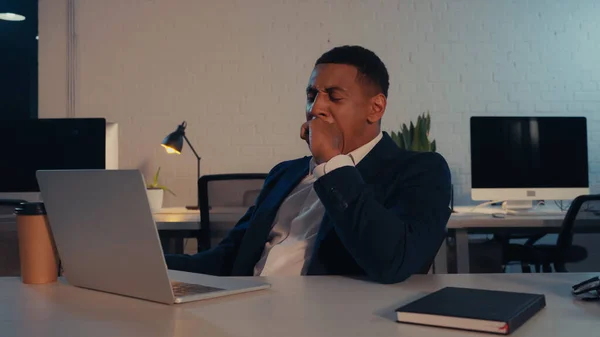 Hombre de negocios afroamericano cansado bostezando cerca de la computadora portátil y la taza de papel en la oficina por la noche - foto de stock