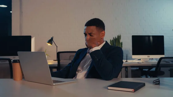 Уставший африканский американский бизнесмен в костюме сидит рядом с ноутбуком и выпивает на вынос в офисе — стоковое фото