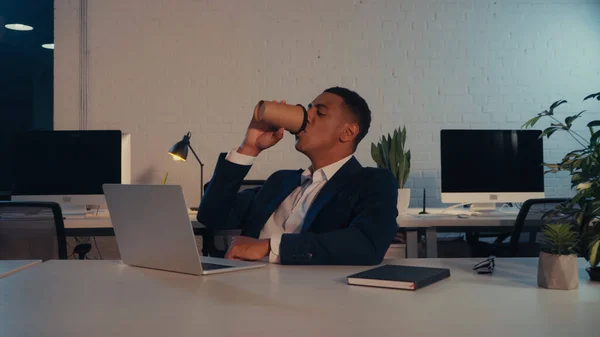 African american businessman drinking coffee to go near laptop in office at night — Stock Photo