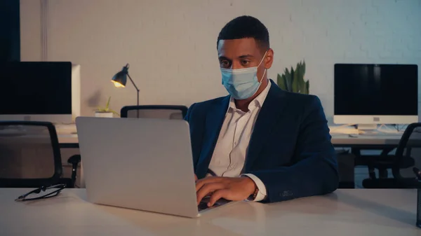 Afroamerikanischer Geschäftsmann in medizinischer Maske mit Laptop nachts im Büro — Stockfoto
