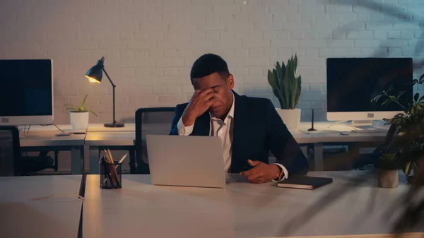 Exhausted african american businessman sitting near laptop and notebook in office at night — Stock Photo