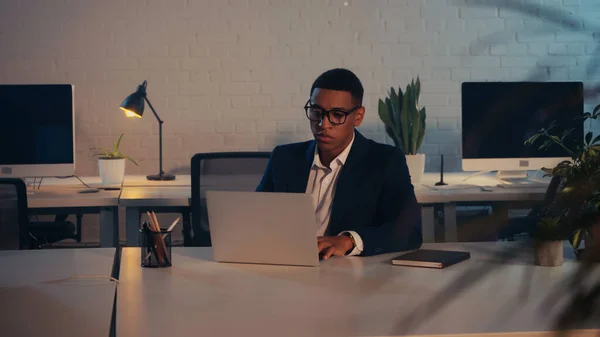 African american manager in eyeglasses using laptop in office at night — Stock Photo