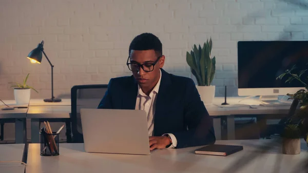 African american manager in suit and eyeglasses using laptop in office at night — Stock Photo