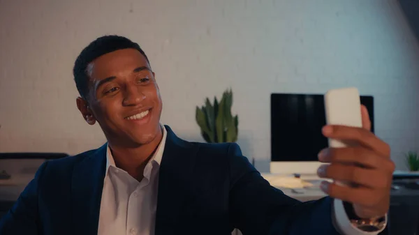 Sonriente hombre de negocios afroamericano tomando selfie en el teléfono inteligente en la oficina por la noche - foto de stock