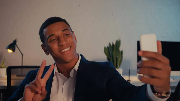 Sonriente hombre de negocios afroamericano tomando selfie y mostrando signo de paz en la oficina por la noche - foto de stock