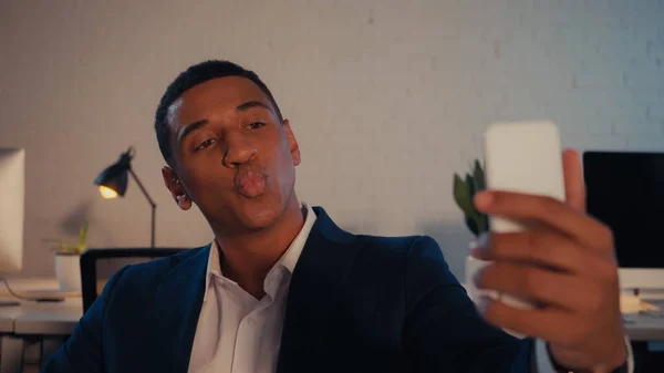 Hombre de negocios afroamericano haciendo pucheros mientras toma selfie en el teléfono inteligente en la oficina - foto de stock