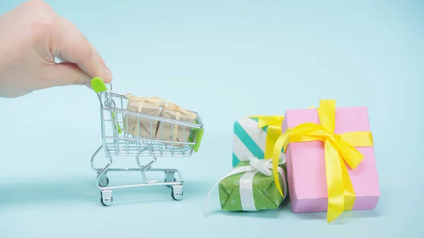 Vista recortada del hombre sosteniendo un pequeño carrito de compras cerca de regalos envueltos en fondo azul - foto de stock