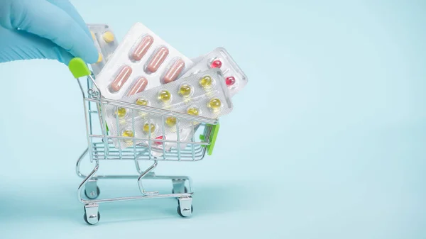 Vista recortada de la persona en guante de látex sosteniendo pequeño carrito de compras con pastillas sobre fondo azul — Stock Photo