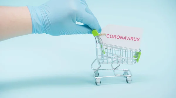 Vista cortada de pessoa em luva de látex segurando carrinho de compras com letras coronavírus no cartão no fundo azul — Fotografia de Stock