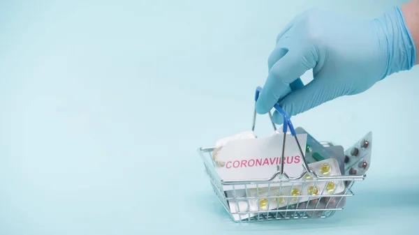 Cropped view of person in latex glove holding shopping basket with coronavirus lettering on card and medicaments on blue — Stock Photo