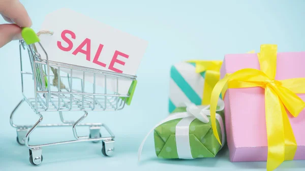 Cropped view of man holding small shopping cart with sale card near wrapped presents on blue background — Stock Photo