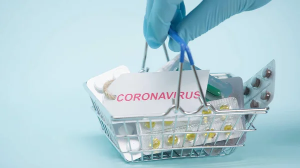 Partial view of person in latex glove holding shopping basket with coronavirus lettering on card and medicaments on blue — Stock Photo