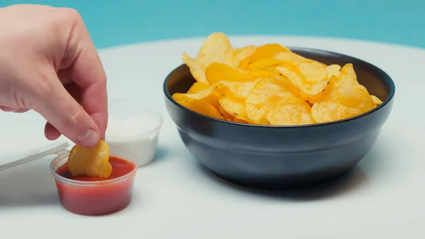 Partial view of man dipping ridged potato chips in ketchup on blue — Stock Photo