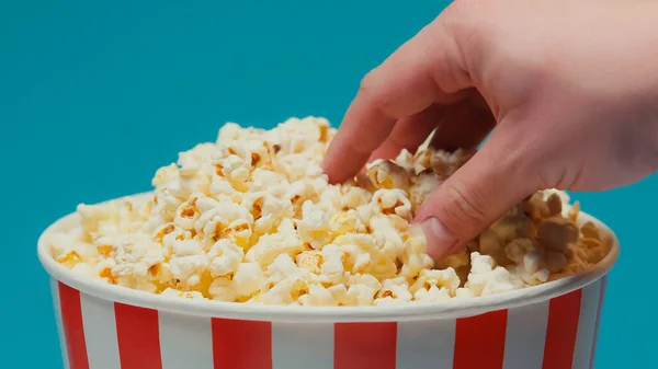 Vista recortada del hombre tomando palomitas de maíz de gran cubo de papel aislado en azul - foto de stock