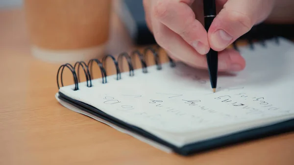 Vista cortada da pessoa segurando caneta enquanto escreve em caderno borrado — Fotografia de Stock