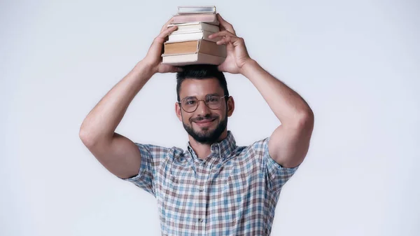 Estudante nerd alegre em óculos segurando pilha de livros acima da cabeça isolado em cinza — Fotografia de Stock