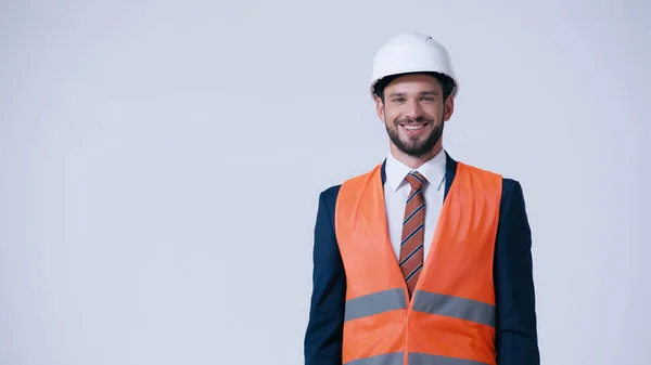 Lächelnder Ingenieur in Bollenhut und Warnweste mit Blick auf Kamera isoliert auf grau — Stockfoto