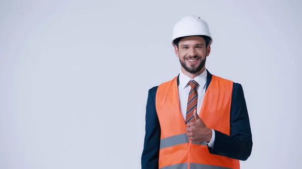 Feliz capataz en chaleco de seguridad y casco mostrando el pulgar hacia arriba aislado en gris - foto de stock