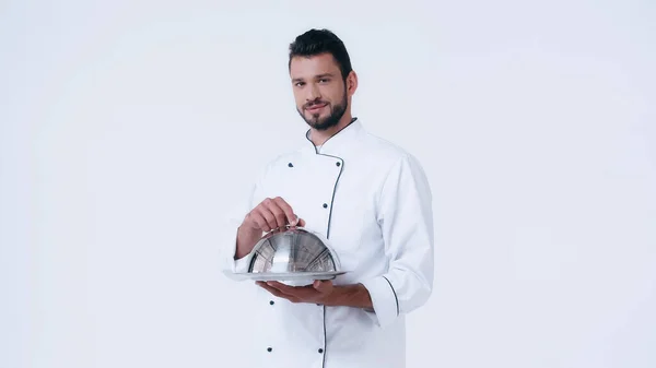 Morena hombre en chaqueta de chef sosteniendo servir plato con cloche y mirando a la cámara aislada en blanco - foto de stock