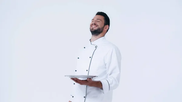 Cheerful chef with closed eyes holding stainless platter isolated on white — Stock Photo