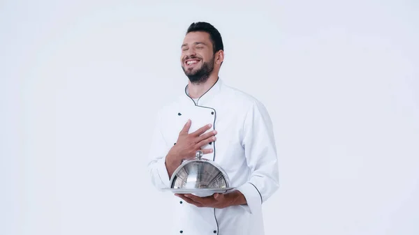 Laughing chef with serving platter and cloche touching chest isolated on white — Stock Photo