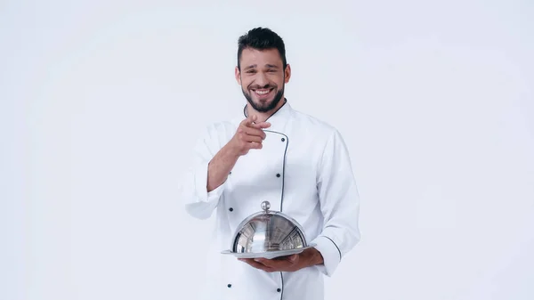 Chef risueño sosteniendo el plato de servir con cloche inoxidable y apuntando con el dedo a la cámara aislada en blanco - foto de stock