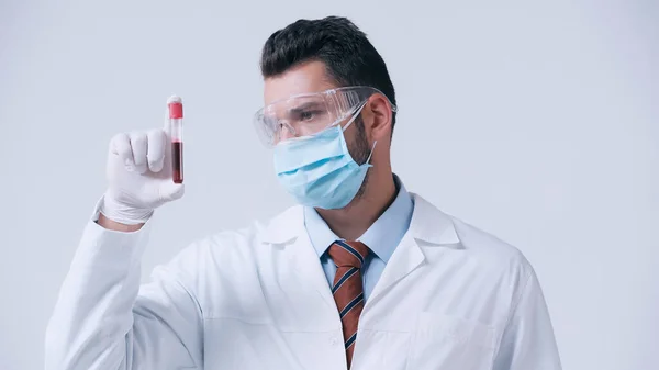 Immunologiste dans le masque médical et les lunettes regardant l'éprouvette avec échantillon de sang isolé sur gris — Photo de stock