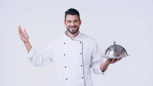 Young chef with serving plate and stainless cloche smiling at camera isolated on white — Stock Photo