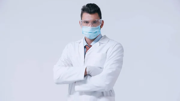Врач в белом халате и медицинской маске с очками, стоящими со скрещенными руками, изолированными на сером — стоковое фото