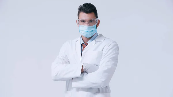 Doctor in white coat and medical mask with goggles standing with crossed arms isolated on grey — Stock Photo