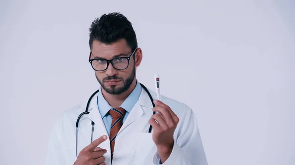 Серьезный врач со стетоскопом, указывающий на электронный термометр, изолированный на сером — стоковое фото