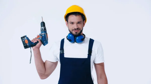 Constructor positivo en hardhat y overoles con taladro eléctrico y mirando a la cámara aislada en blanco - foto de stock