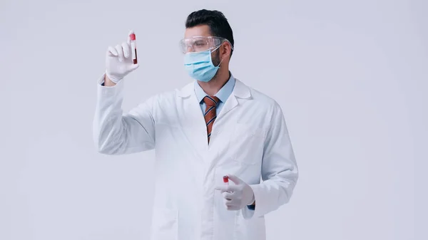 Doctor in white coat and medical mask with goggles holding test tubes isolated on grey — Stock Photo