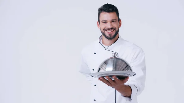 Smiling chef standing with hand behind back while holding serving dish with cloche isolated on white — Stock Photo