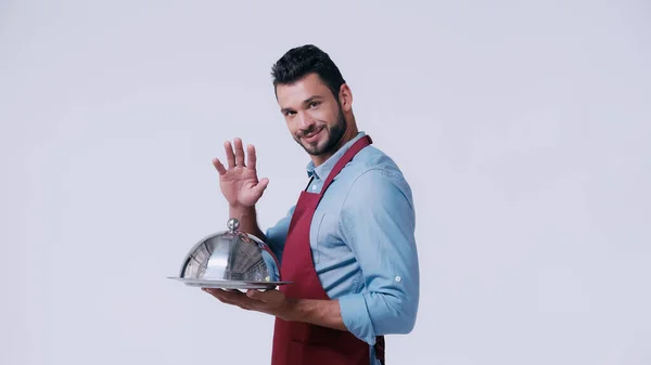 Positive waiter waving hand while holding serving dish with cloche isolated on grey — Stock Photo